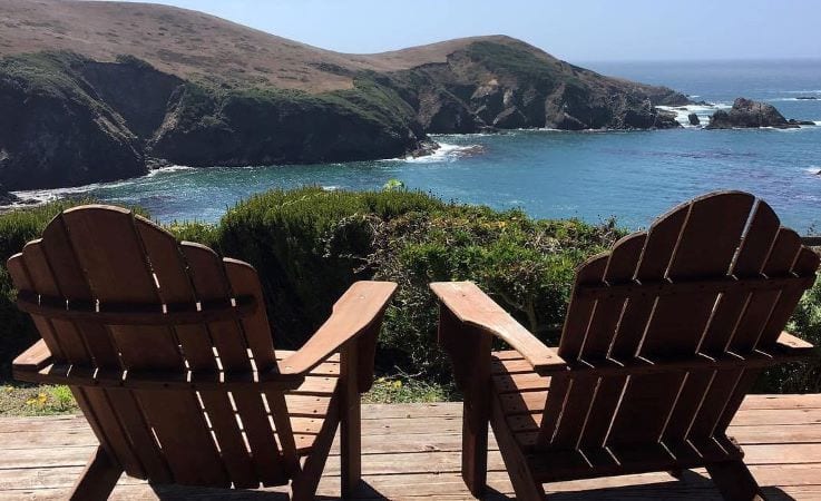 A pair of adirondack chairs are set up beside each other on a wooden deck overlooking the California coast. Waves crash into rocky bluffs, and the water is dark blue.