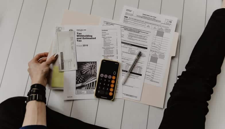 A person is sitting on the floor with tax forms laid out in front of them, and the calculator app open on their cell phone. 