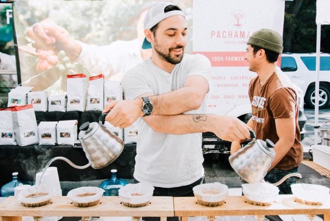 A Californian barista brews coffee at a pop-up coffee stand. He has six coffee-filled filters lined up in custom trays in front of his and he’s impressively using two gooseneck kettles at the same time to pour hot water into the filters. 