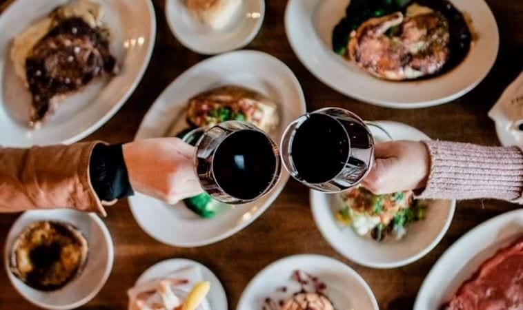 Overhead view of the hands of two women clinking their wine glasses together over a feast of entrees and appetizers at a restaurant in California.