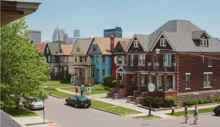 Locals walk through Detroit’s Corktown neighborhood on a sunny day. The neighborhood is filled with large, two-story homes, and the city’s skyline is visible in the distance.