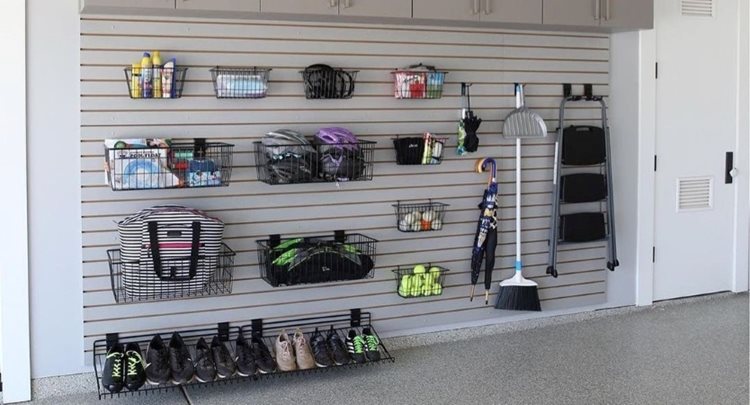A garage wall that has horizontal slats installed to allow for hooks and baskets to easily hang from anywhere. Most of the items being stored on the wall are sports-related, such as balls, shoes, and other gear. 