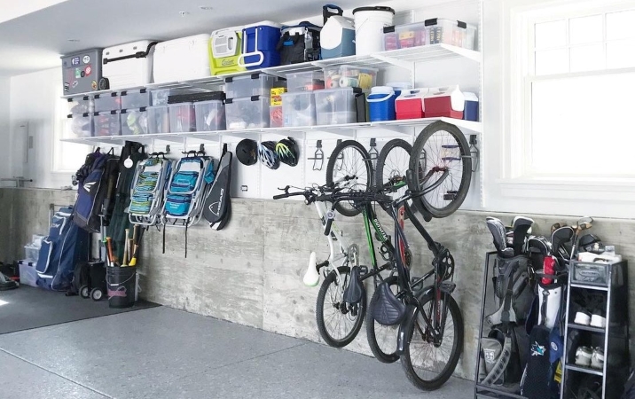 A garage wall with shelves installed high on the wall. Plastic bins and coolers line the shelves, and larger items — like bicycles, lawn chairs, and sports equipment — hang below the shelves to make the most of the space. 