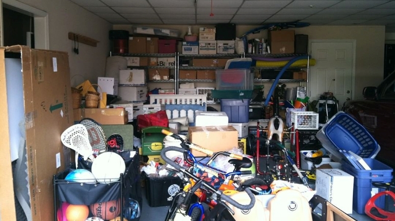 A very cluttered residential garage that is in desperate need of DIY garage organization. There are shelves in the back of the garage, but they’re filled up with boxes and other random items. The floor space is entirely covered in a large pile of various items, from plastic baskets and a large toy horse to bicycles and sports equipment. 