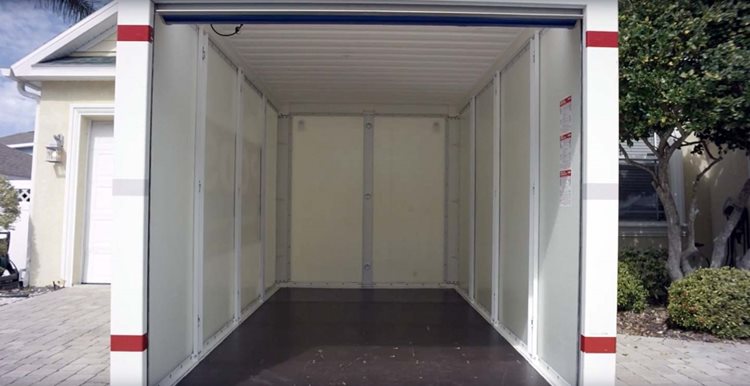 View of the inside of an empty PODS portable storage container. The interior walls of the container are white, and the floor is dark gray. The container is sitting in the driveway of a residential home. 