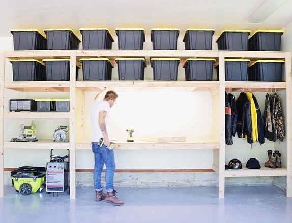 A man inspects his work after building his own DIY garage shelves to organize his garage. The shelves are made with 2” x 4” wooden beams and have already been filled with neatly arranged black and yellow plastic bins, various power tools, some work jackets, helmets, and boots. 