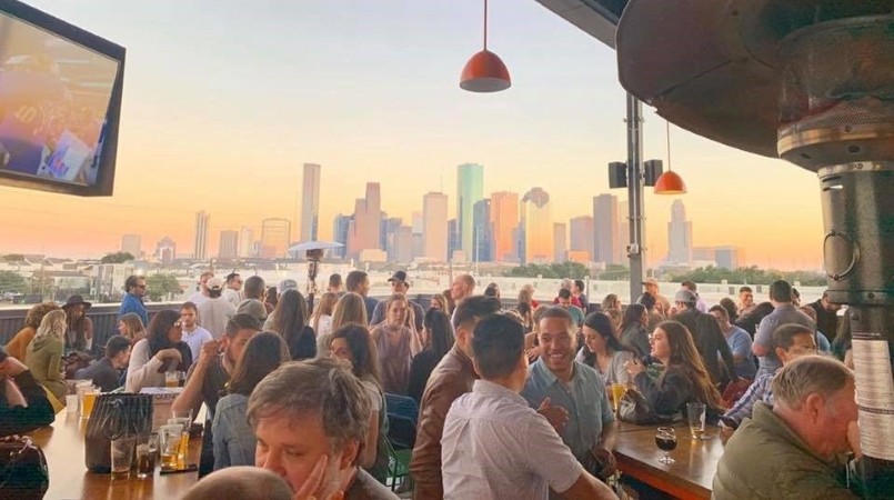 Dozens of people enjoy Houston nightlife at a rooftop bar as the sun sets behind the city skyline.