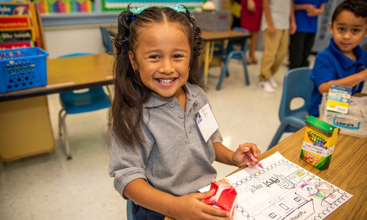 A young student in Houston, Texas, smiles while coloring at school. Another student is working on his own artwork beside her.