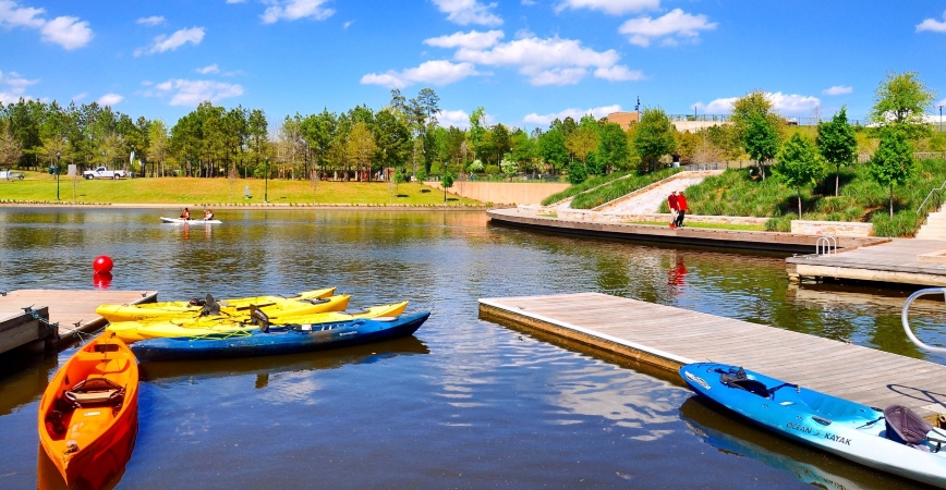 People walk around a lake on a sunny day in The Woodlands, one of the best Houston suburbs. There are a few docks with kayaks floating beside them, ready to be taken out into the water. 