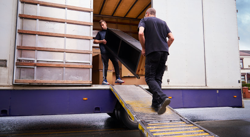 Two professional movers from a full-service moving company carry a piece of hard-wood furniture up a ramp and into a moving truck. 
