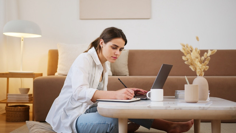 A young woman is sitting on her living room floor, using her laptop to research moving companies online, and taking notes on paper.