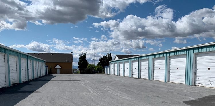 A view of two rows of outdoor self-storage units under a blue sky filled with white clouds. Each storage unit features a garage-style door. 