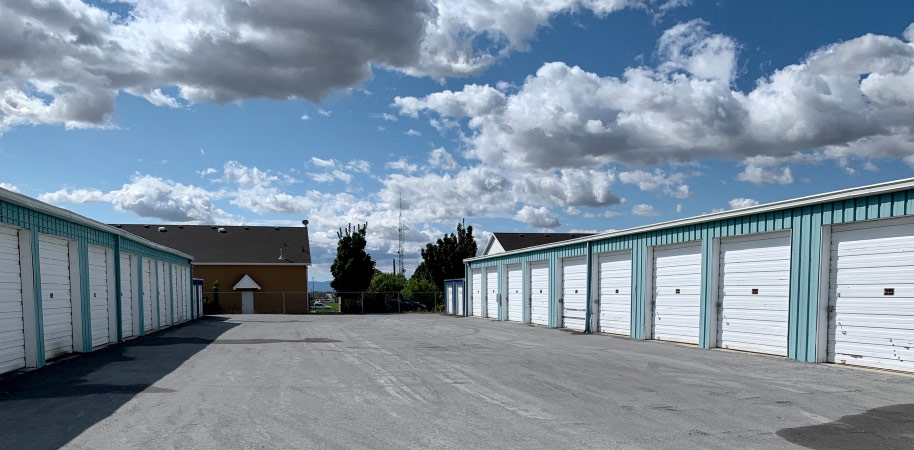 A view of two rows of outdoor self-storage units under a blue sky filled with white clouds. Each storage unit features a garage-style door. 