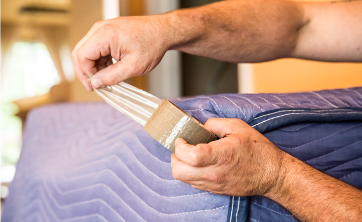 Close-up view of a man using packing tape to secure a blue packing blanket around the corner of a piece of furniture. 