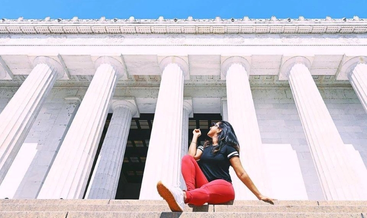 A woman in a black shirt and red pants is sitting atop the steps of The Lincoln Memorial, looking and pointing to the monument behind her.