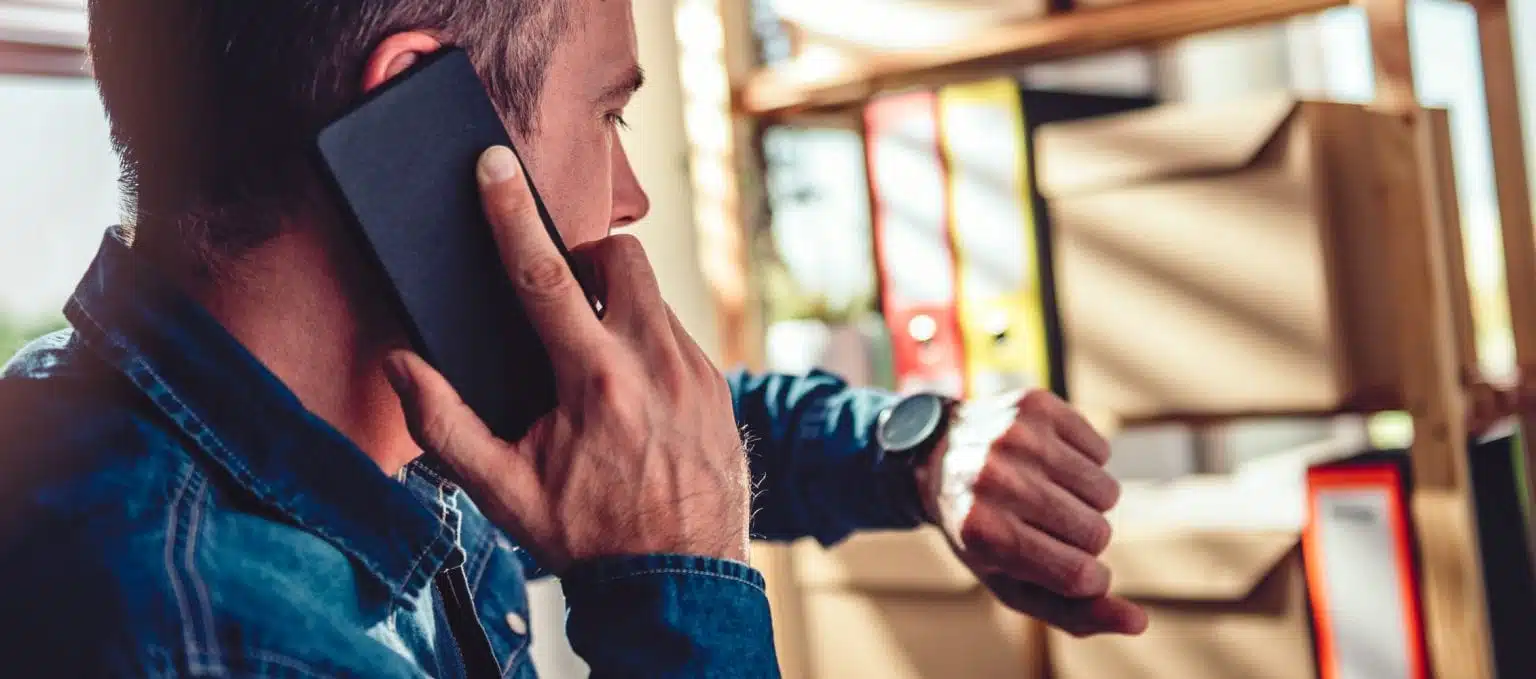 A man checks his watch while he's on the phone. He’s in the middle of a last-minute move and there are moving boxes visible in the background.