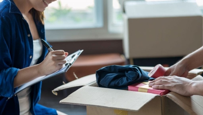 A woman making a moving checklist as another person packs a moving box. 