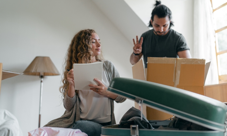 A woman is noting down what is being packed for an upcoming move as her husband looks through a moving box, indicating how many of a particular item is inside the box.