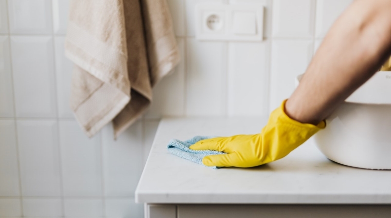 Close-up view of a hand in a yellow rubber glove, wiping off a bathroom counter. 