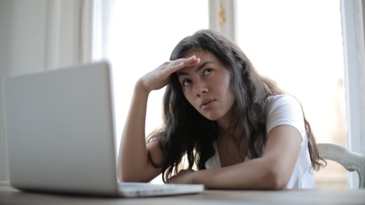 A young woman is sitting in front of her laptop with her hand on her head, looking up and thinking.