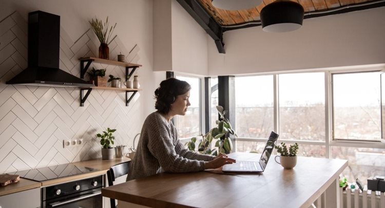 A young woman is sitting at her kitchen island, using her laptop to do online research into Airbnb hosting.