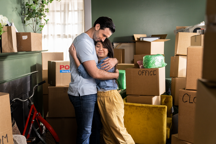 A couple embraces in the middle of the living room of their new Charlotte, NC, home. It's filled with moving boxes and furniture.