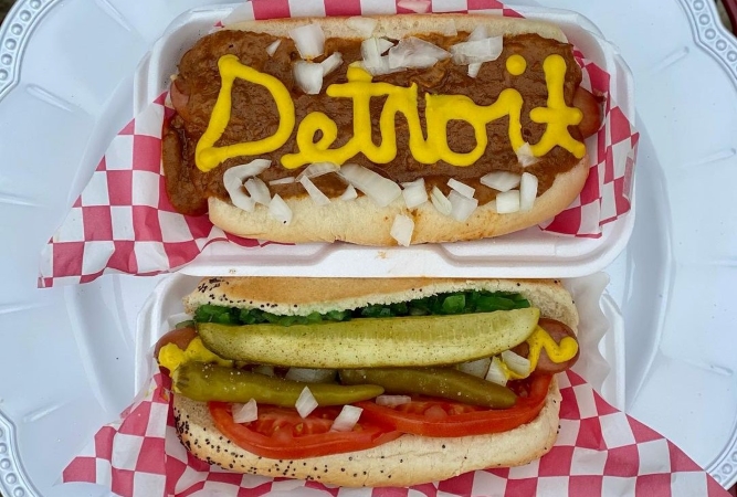A chili hot dog with the word “Detroit” written on it in mustard and another hot dog with all the fixings. 