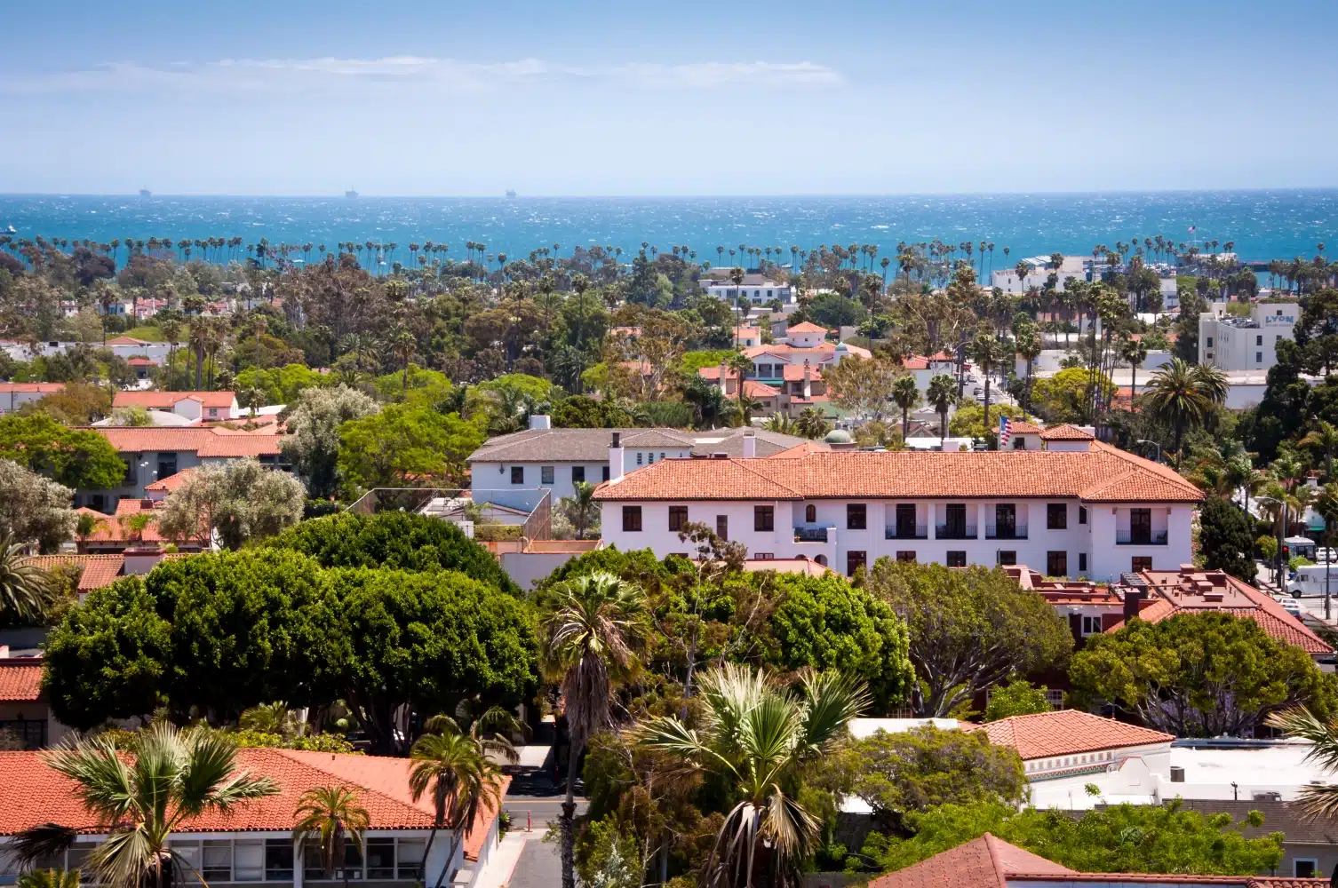 Treetop view of Downtown Santa Barbara, California, on a sunny day. Many of the buildings feature a white stucco exterior and red tile roofs. In the distance, the Pacific Ocean is visible. 