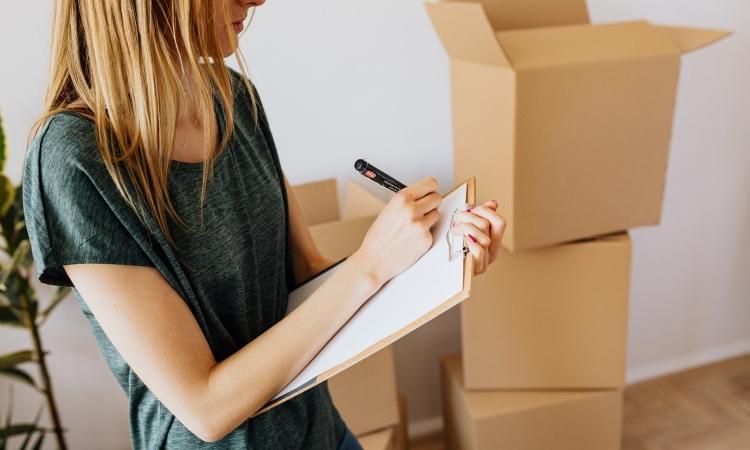 A woman is using a clipboard to hold her packing list as she notes down what she plans to pack for her move to Santa Barbara, California. On the floor behind her are two stacks of moving boxes ready to be packed. 