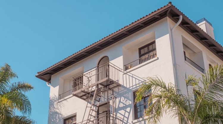 View from below of the top half of an apartment building in Downtown Santa Barbara. The building features a white stucco exterior and rile roof. There’s a fire escape and a small balcony on the side of the building.