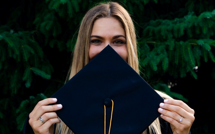 A young woman is smiling behind her graduation cap, which she’s holding in front of her face. She’s a student of the University of California at Santa Barbara.