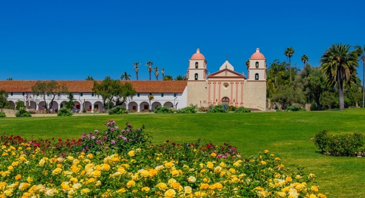 Distant view of Old Mission Santa Barbara from across a green field. The Mission is an old Spanish-style building with white stucco archways. In the foreground, a patch of colorful flowers is blooming.