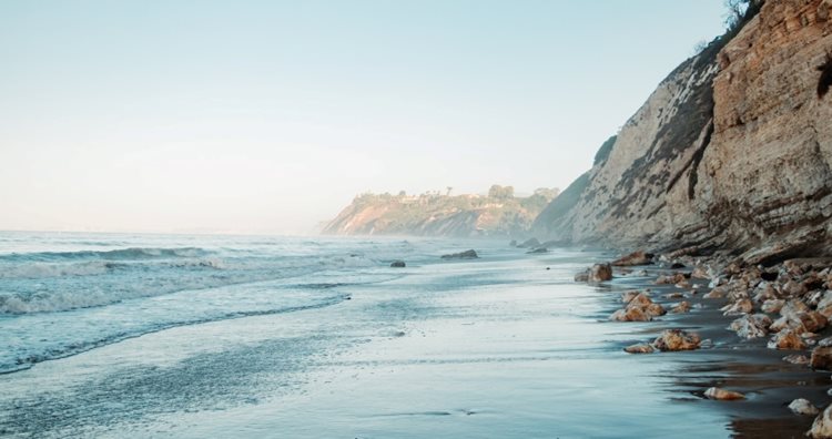 View of the Santa Barbara bluffs from the beach in The Mesa. The bluffs feature steep rocky walls and the sand is a dark brown.