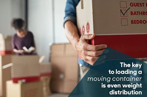 Close-up of a man carrying a moving box while a woman looks through paperwork in the background. There are several moving boxes in the room.