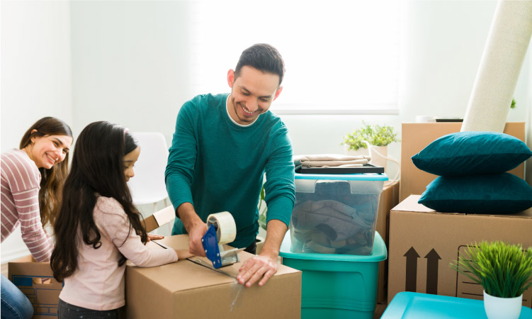 A young girl is helping her father tape up a moving box as her mother packs another box beside them. The room is filled with packed moving boxes, plastic tubs, and other miscellaneous home goods.