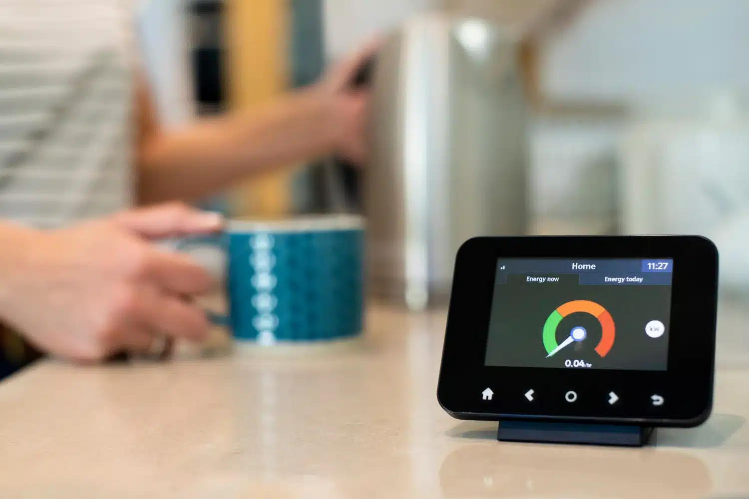 A small home energy meter sits on a kitchen island. The display appears to show a good energy reading. Out of focus in the background, a person is holding a mug and a coffee carafe, preparing to pour a cup of coffee.