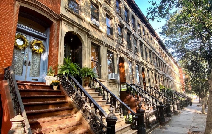 View of a row of townhomes in Jersey City, New Jersey. They look very similar to one another, with stone or brick exteriors, steep entryway stairs, and wrought-iron railings. 