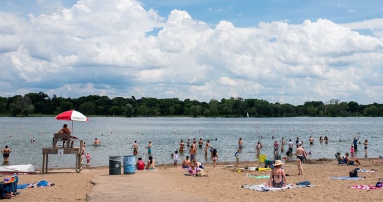 Dozens of locals enjoy good weather at one of the many lakes in Minneapolis, Minnesota. A lifeguard sits on a stand under a red and white umbrella as visitors swim and sunbathe on the sandy beach of the lake. 
