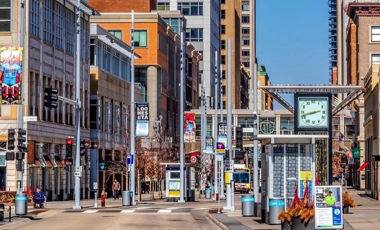 Street-level view of an intersection in downtown Minneapolis. There are commercial buildings and a bus stop with a large clock face on display. 