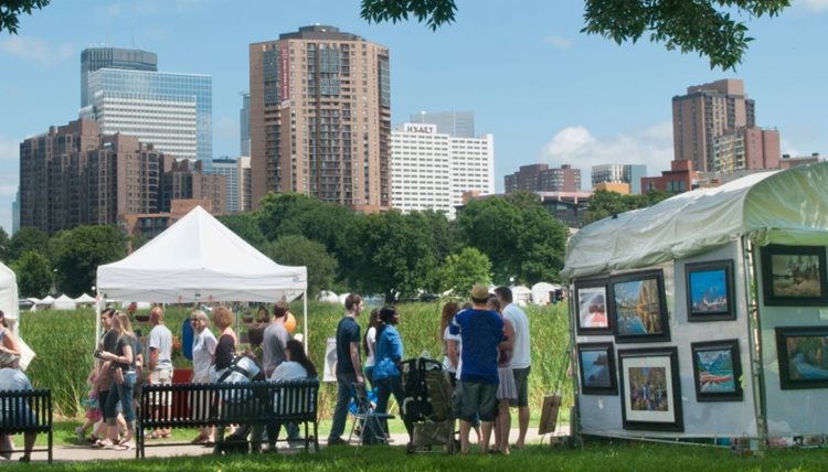 Locals stroll through the Loring Park Art Festival in Minneapolis, Minnesota. There are several white tents set up throughout the park where vendors display their wares, and the city skyline can be seen in the distance. 