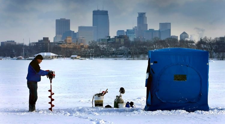 A man gets ready to do some ice fishing with the Minneapolis skyline in the background. He has a blue box tent with a collection of fishing equipment arranged nearby, and he’s using a power tool to drill a hole in the ice.