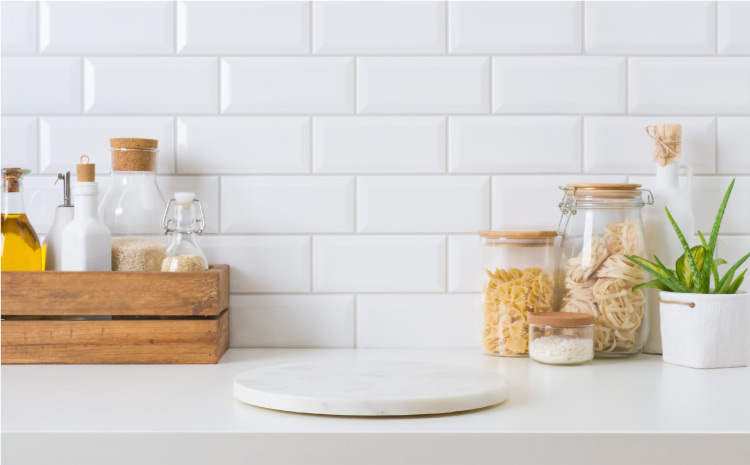 Close-up view of a white kitchen countertop. Decorative glass bottles and jars of dry goods and cooking oils are arranged neatly against the subway tile backsplash or in an open wooden box. There’s a white lazy Susan on the counter as well.