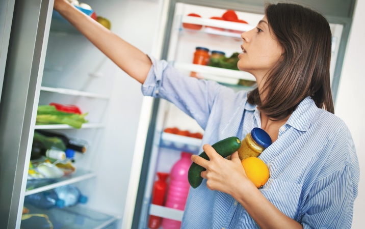 A young woman is holding an orange, a cucumber, and a jar of peanut butter in one hand, as she uses her other hand to reorganize the inside of her fridge.