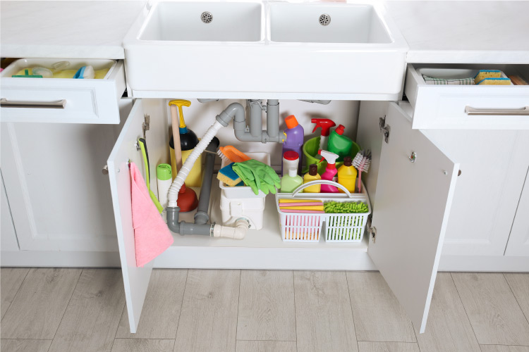 The space under a kitchen sink, neatly organized with plastic trays and baskets to hold cleaning supplies, and small hooks on the door to hold rags. 