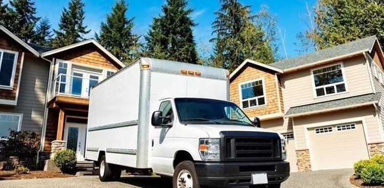 A white box moving truck is parked in a residential driveway with the back of the truck facing the front of the house. 