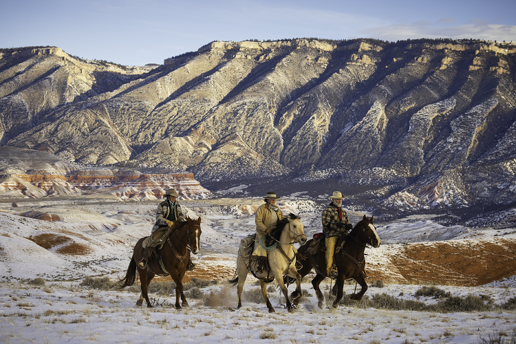 Two men and a woman on horseback on a snowy Wyoming ranch. Mountains tower in the background
