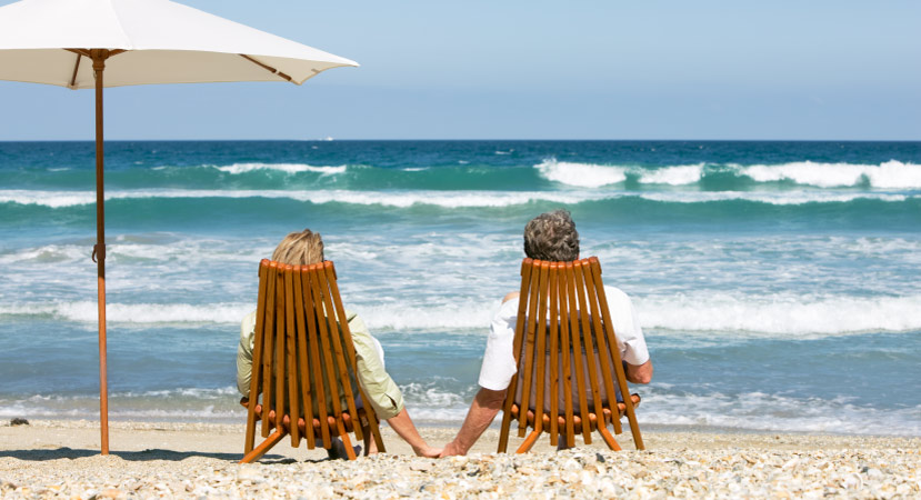 A retired couple lounges on two wooden chairs on a beach in Florida. They’re holding hands and looking out at the surf of the blue Gulf waters.