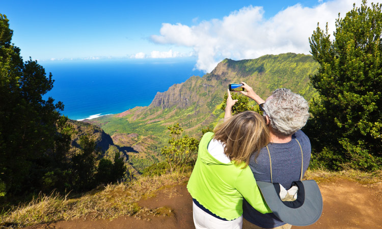 A couple is taking a photo of the scenic view from the Kalalau Lookout in the Waimea Canyon of Kauai, Hawaii. The sky and sea are both vibrant blue, and fluffy white clouds are moving in from over the mountains.