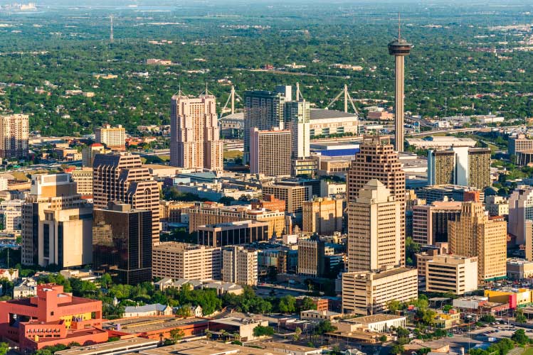Aerial view of the city of San Antonio, Texas. It’s a sunny, summer day and the tall buildings of Downtown San Antonio are shining brightly. In the distance, flat suburban neighborhoods are lush with mature, green trees.