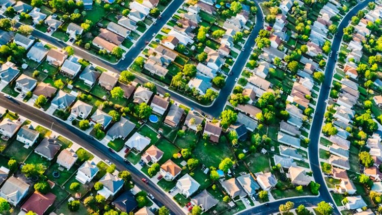 A birds-eye view of a residential suburban neighborhood outside San Antonio, TX. Large single-family homes are neatly arranged along curving suburban streets. Every backyard is filled with dark green grass and large trees.